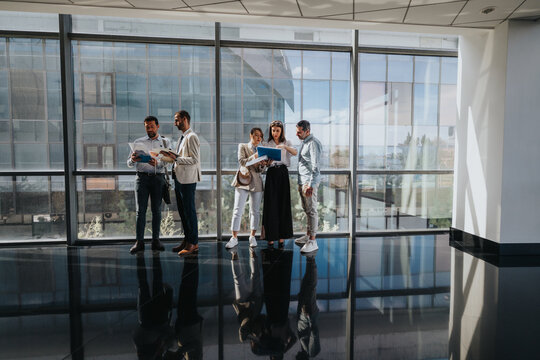 一支多元化的专业团队站在明亮的玻璃幕墙中庭，集体审阅文件和文件夹，在现代企业环境中协作交流、分享想法