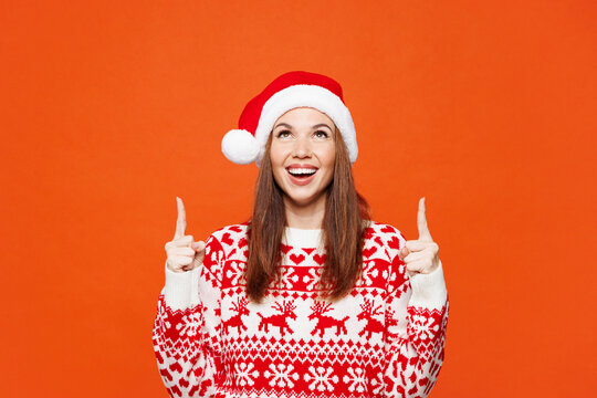 快乐的年轻女子身着红色保暖圣诞毛衣，头戴圣诞帽，摆出姿势，用食指指向头顶上方的空白区域模型，背景为纯橙色。新年快乐庆祝节日主题
