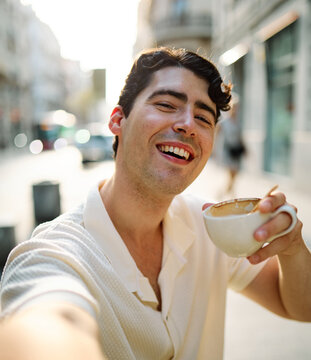 Portrait of a smiling young man guy with a coffee cup in a coffee shop or a restaurant in the city, tourists visiting destination, summer trip exploring, posing for a selfie photo with mobile phone